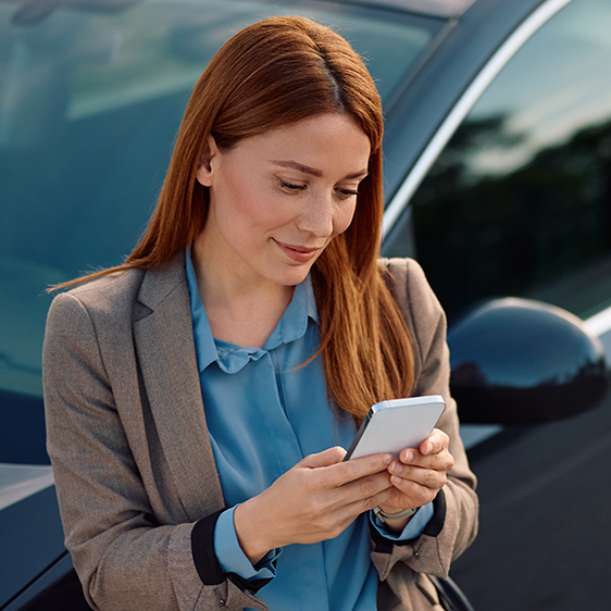 Happy businesswoman using cell phone while standing beside her car on parking lot.